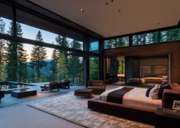 Luxurious bedroom with floor-to-ceiling windows showcasing a forest view at sunset. Features a large bed, cozy chairs, and warm, elegant decor.