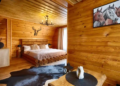 A cozy wooden cabin bedroom with a rustic log bed, deer antler decor, and horse painting. A cowhide rug adds warmth; soft light filters through curtains.