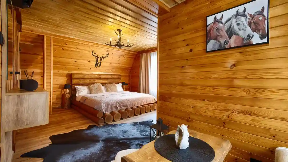 A cozy wooden cabin bedroom with a rustic log bed, deer antler decor, and horse painting. A cowhide rug adds warmth; soft light filters through curtains.