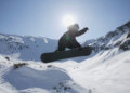 A snowboarder performs a high jump against a bright sunlit sky in a snowy mountain landscape, capturing a thrilling and adventurous moment.