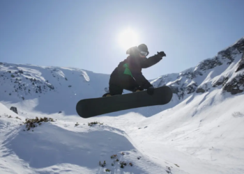 A snowboarder performs a high jump against a bright sunlit sky in a snowy mountain landscape, capturing a thrilling and adventurous moment.