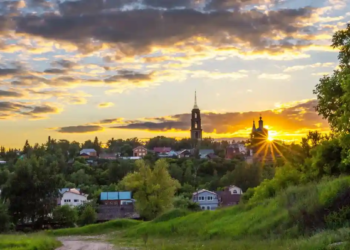 Sunset over a tranquil village with lush greenery and scattered houses, a church tower silhouetted against a vibrant sky, evoking serenity.