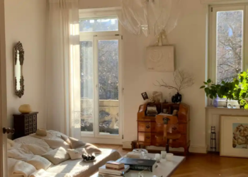 Sunlit bedroom with a low bed, soft pillows, and a cozy, inviting atmosphere. Wooden dresser, potted plant, and large windows add warmth and charm.