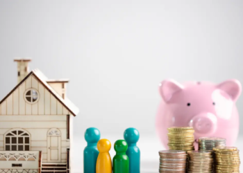 A miniature house and colorful pegs stand beside a pink piggy bank and stacks of coins, symbolizing family savings and financial planning.