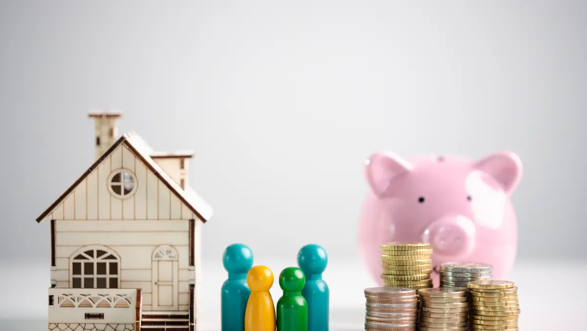 A miniature house and colorful pegs stand beside a pink piggy bank and stacks of coins, symbolizing family savings and financial planning.