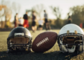 Two football helmets are on turf next to a football