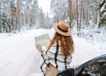 A woman in a brown hat and plaid shawl studies a map next to a car on a snowy forest road. The scene feels adventurous and serene.