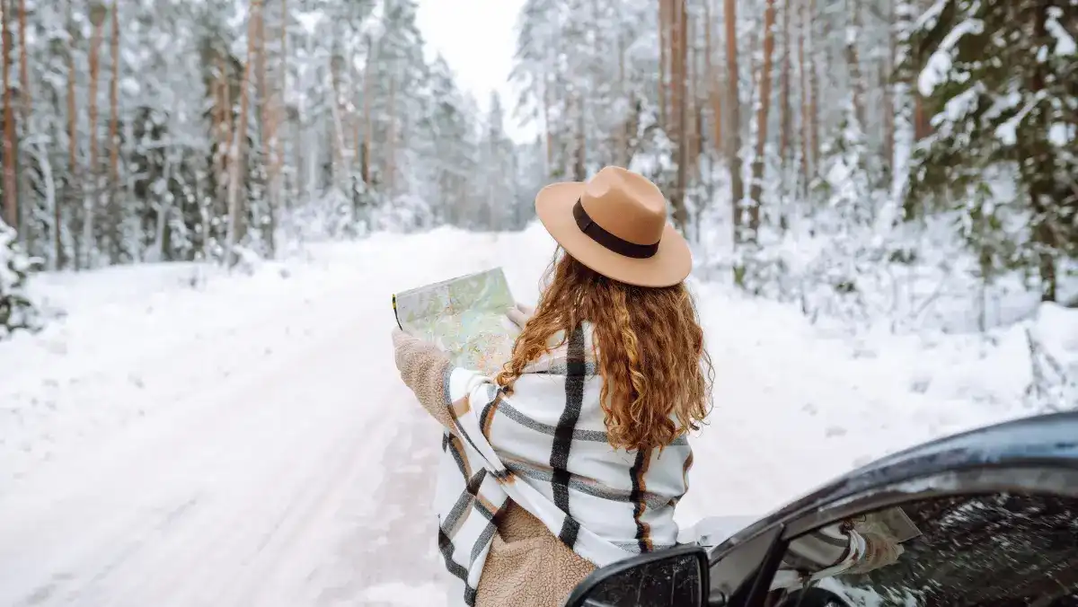 A woman in a brown hat and plaid shawl studies a map next to a car on a snowy forest road. The scene feels adventurous and serene.