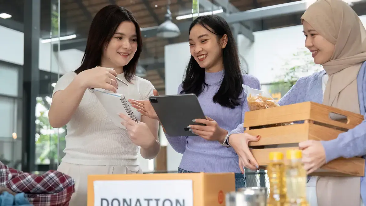 Three women happily organize donations in a modern setting. One holds a notebook, another a tablet, and the third carries a box. A donation box is visible.