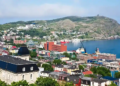 Colorful buildings nestled by a calm bay in St. John's, Newfoundland, with green hills and blue skies, conveying a vibrant and serene coastal scene.
