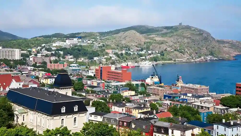 Colorful buildings nestled by a calm bay in St. John's, Newfoundland, with green hills and blue skies, conveying a vibrant and serene coastal scene.