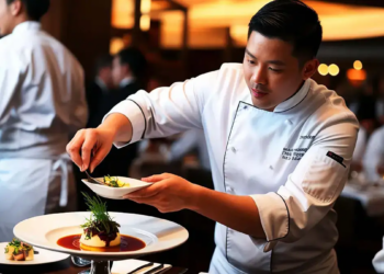 A focused chef in a white uniform artfully garnishes a dish with herbs in an elegant restaurant kitchen, surrounded by busy staff, creating a lively atmosphere.