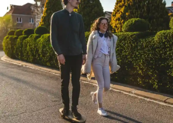 A man on a skateboard and a woman walking beside him smile while passing trimmed bushes on a sunny street, conveying a cheerful, relaxed moment.