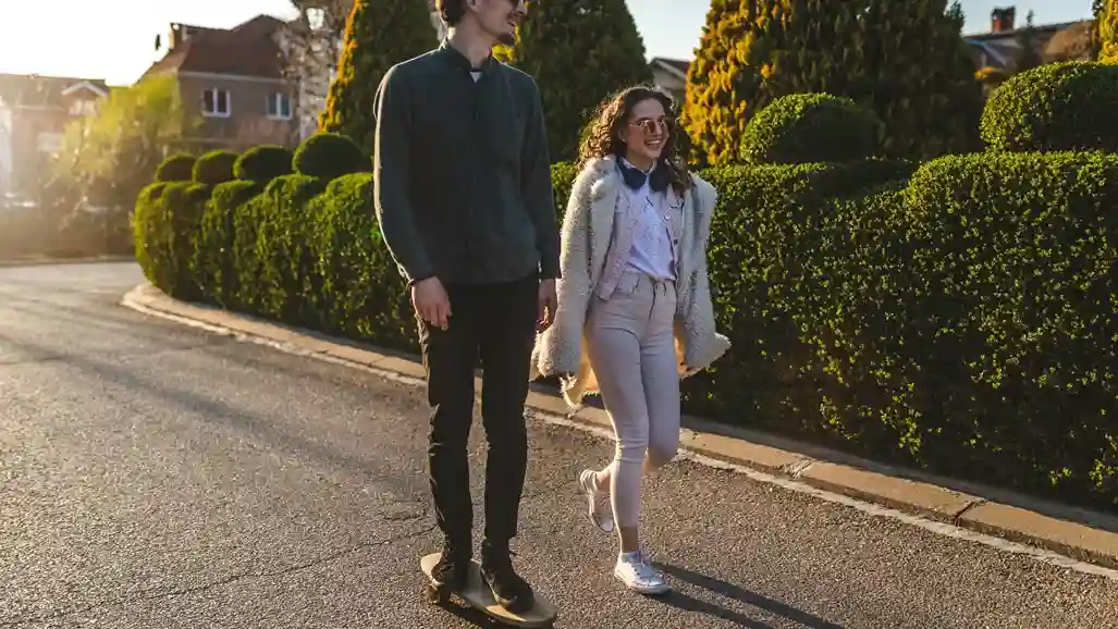 A man on a skateboard and a woman walking beside him smile while passing trimmed bushes on a sunny street, conveying a cheerful, relaxed moment.