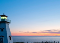 A white lighthouse with a glowing lantern stands against a vibrant sunset sky, overlooking calm ocean waters. The scene is serene and picturesque.