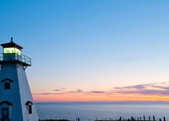A white lighthouse with a glowing lantern stands against a vibrant sunset sky, overlooking calm ocean waters. The scene is serene and picturesque.