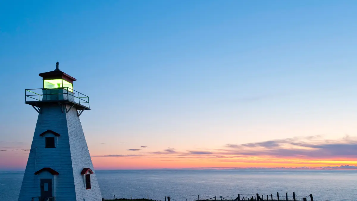 A white lighthouse with a glowing lantern stands against a vibrant sunset sky, overlooking calm ocean waters. The scene is serene and picturesque.