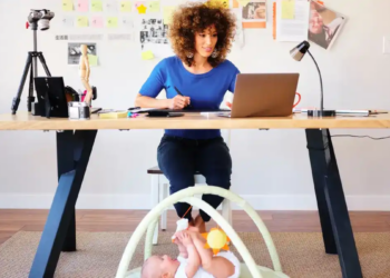 A woman works at a desk, focused on her laptop, surrounded by notes and tools. Below, a baby plays on a mat. The scene is busy yet warm.