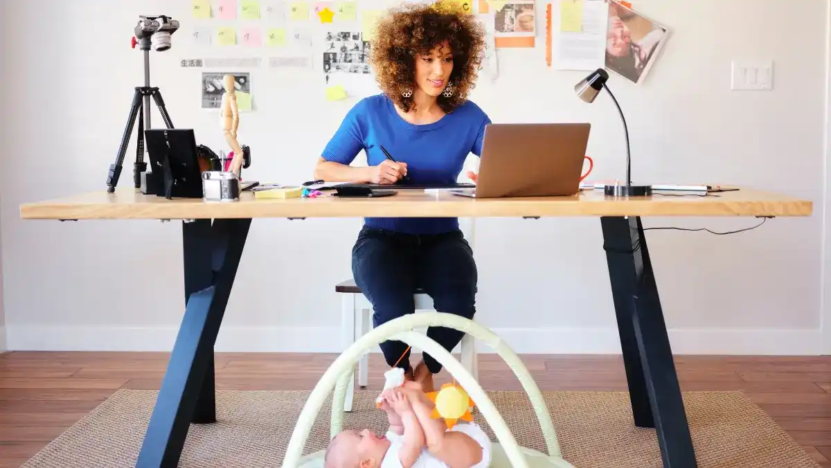 A woman works at a desk, focused on her laptop, surrounded by notes and tools. Below, a baby plays on a mat. The scene is busy yet warm.
