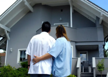 A couple stands arm in arm, gazing at a two-story gray house with a white roof.