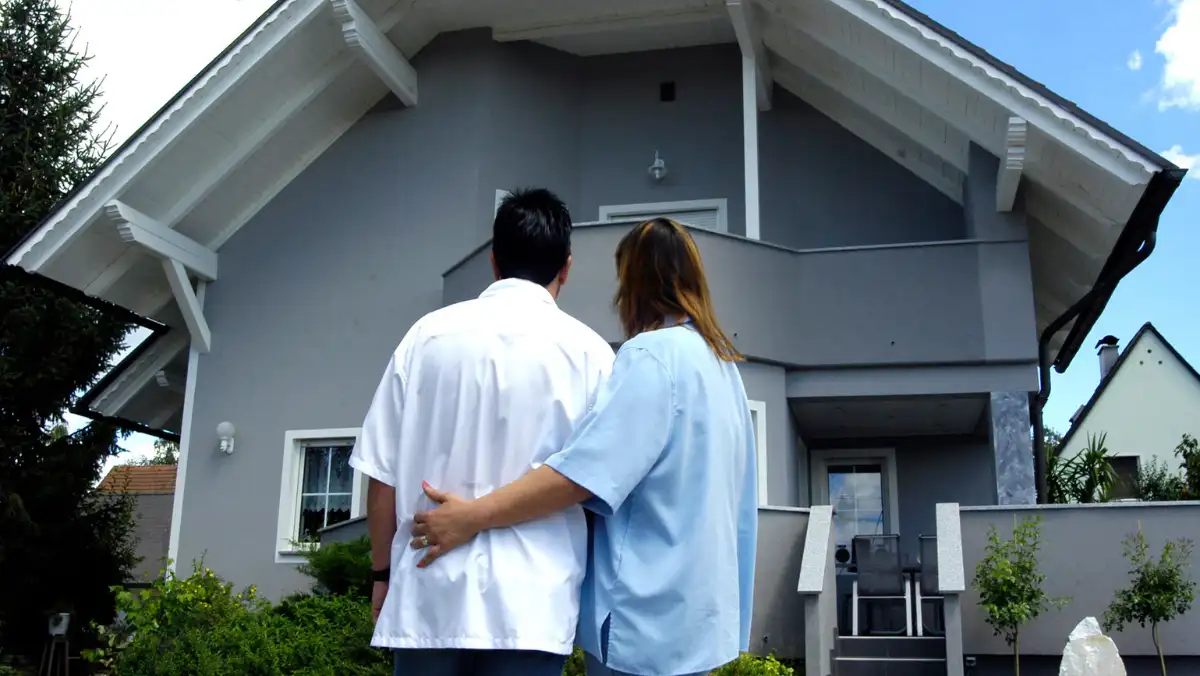 A couple stands arm in arm, gazing at a two-story gray house with a white roof.
