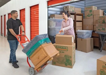 “A man pushes a dolly with nightstand on it toward his storage unit. A woman stands in front of the storage unit taping a moving box.”/>