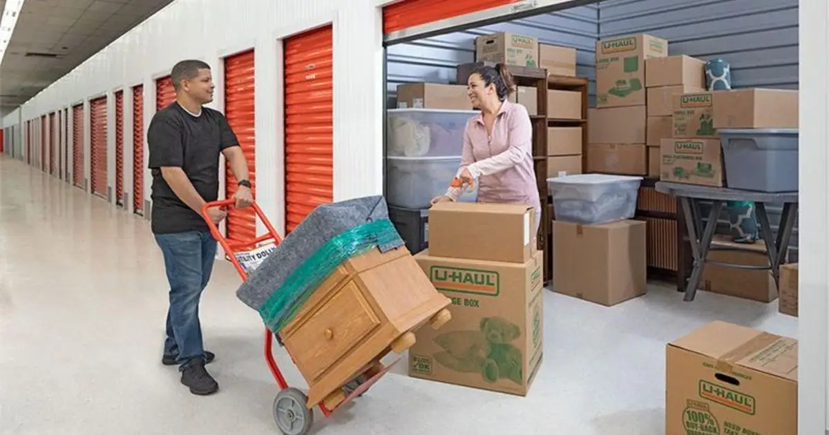 “A man pushes a dolly with nightstand on it toward his storage unit. A woman stands in front of the storage unit taping a moving box.”/>
