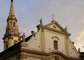 "Baroque church façade with ornate spire and statues at sunset. The building is cream-colored with decorative details, conveying a serene and historic atmosphere."