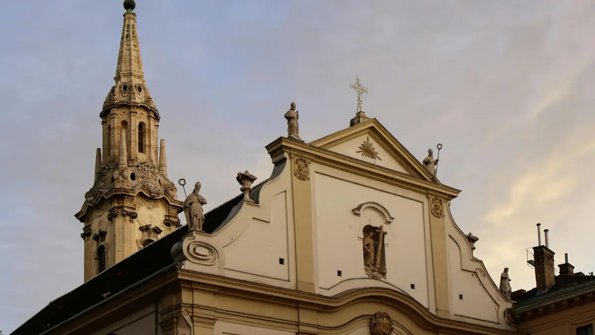 "Baroque church façade with ornate spire and statues at sunset. The building is cream-colored with decorative details, conveying a serene and historic atmosphere."