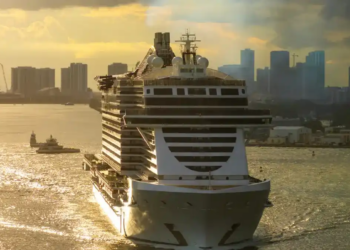 A large cruise ship sails towards a city skyline at sunset. The warm golden light reflects off the water, creating a serene and majestic atmosphere.