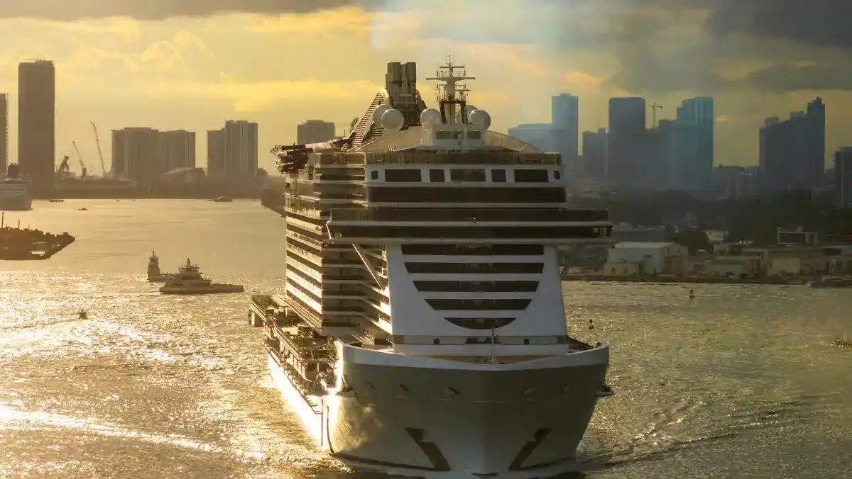 A large cruise ship sails towards a city skyline at sunset. The warm golden light reflects off the water, creating a serene and majestic atmosphere.