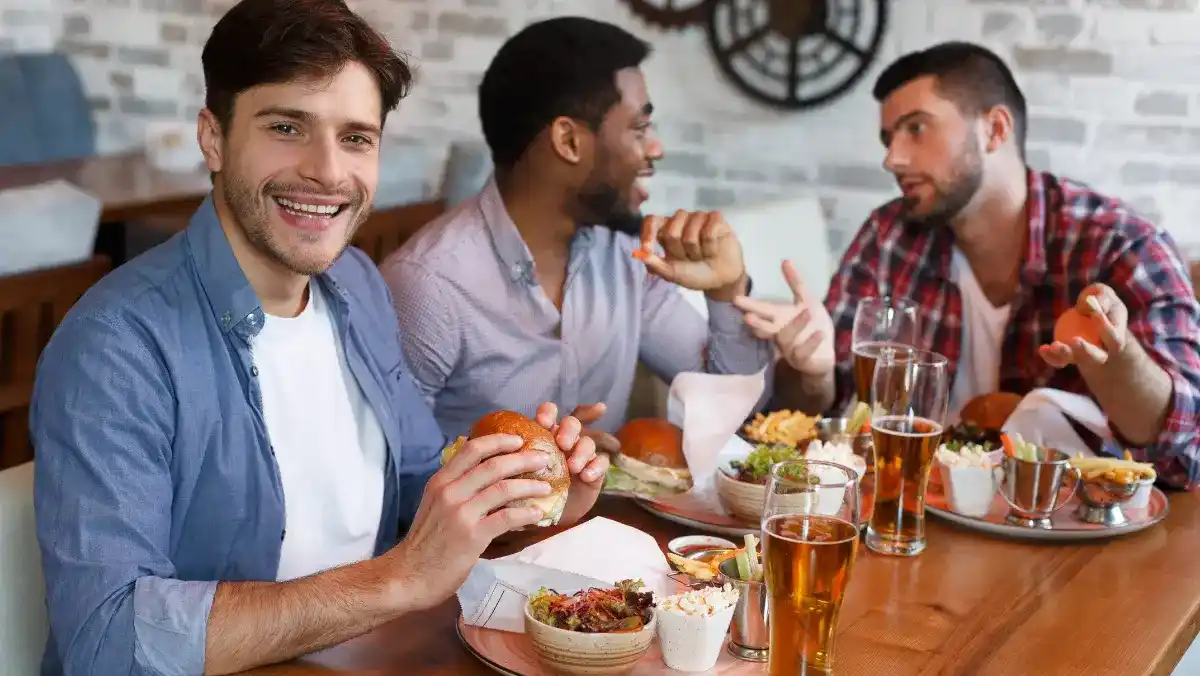Three friends enjoying a meal at a restaurant, laughing and chatting. Burgers, fries, and beer are on the table. The atmosphere is lively and cheerful.