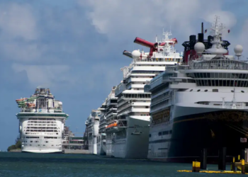 Several large cruise ships docked at a port under a partly cloudy sky. The ships are lined up side by side, creating a sense of grandeur and anticipation.