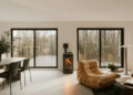 Modern living room with large windows overlooking a forest. Features a cozy fireplace, tan lounge chairs, a sleek dining set, and soft lighting.