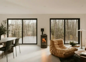 Modern living room with large windows overlooking a forest. Features a cozy fireplace, tan lounge chairs, a sleek dining set, and soft lighting.