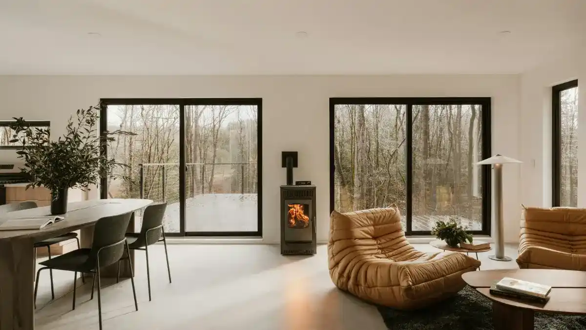 Modern living room with large windows overlooking a forest. Features a cozy fireplace, tan lounge chairs, a sleek dining set, and soft lighting.