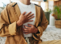 A person sits cross-legged indoors, wearing a tan corduroy outfit. They place one hand on their chest and the other on their stomach, suggesting calmness and mindfulness.