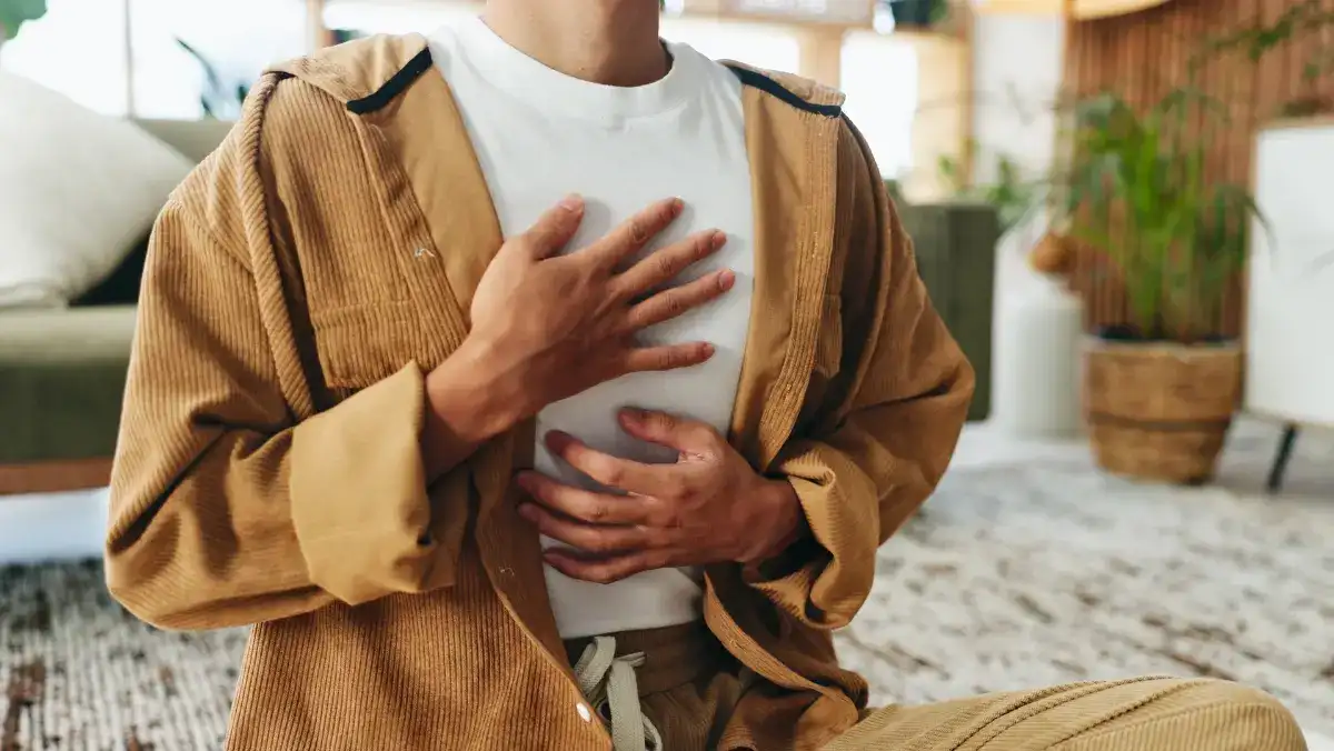 A person sits cross-legged indoors, wearing a tan corduroy outfit. They place one hand on their chest and the other on their stomach, suggesting calmness and mindfulness.