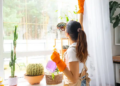 A woman wearing orange gloves and an apron cleans a large window in a bright room filled with various houseplants. The scene conveys a sense of freshness and productivity.