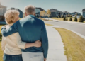 Elderly couple walks arm in arm on a suburban street under a clear blue sky, conveying warmth and companionship. Houses and manicured lawns line the path.