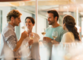 Four people enjoy a wine tasting, holding glasses and smiling in a bright, modern setting. The mood is joyful and social, with sunlight streaming in.