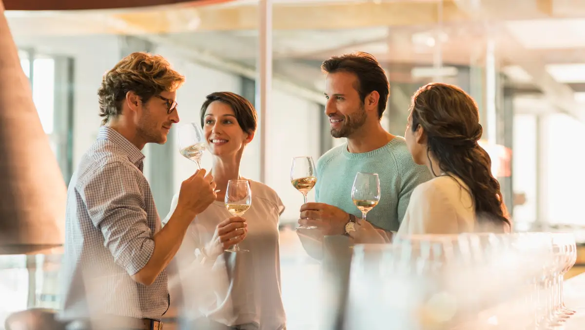 Four people enjoy a wine tasting, holding glasses and smiling in a bright, modern setting. The mood is joyful and social, with sunlight streaming in.