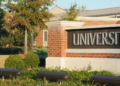 A brick wall with "UNIVERSITY" in large silver letters, surrounded by lush greenery and trees in soft daylight, creating a welcoming academic setting.