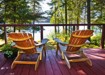 Two wooden chairs sit on a red deck overlooking a serene lake. Surrounded by lush trees, the scene conveys tranquility and natural beauty.