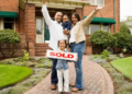 A joyful family of four poses outside their new brick house. The father and mother raise their arms in celebration, while the daughter holds a "Sold" sign.