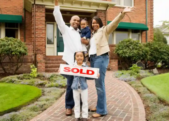 A joyful family of four poses outside their new brick house. The father and mother raise their arms in celebration, while the daughter holds a "Sold" sign.
