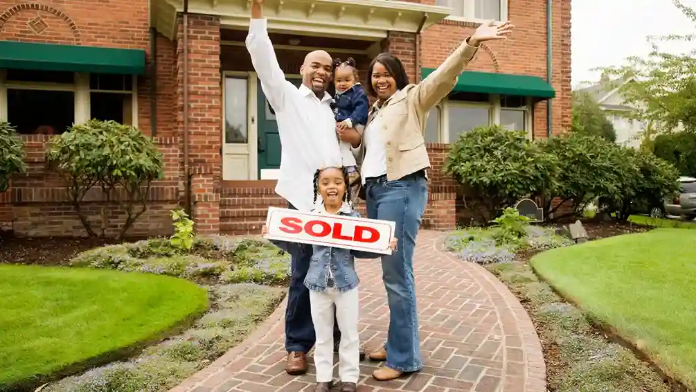 A joyful family of four poses outside their new brick house. The father and mother raise their arms in celebration, while the daughter holds a "Sold" sign.