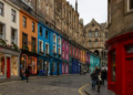 Vibrant street with colorful shopfronts in shades of orange, blue, pink, and red. Stone buildings and cobblestone road enhance the historic feel.
