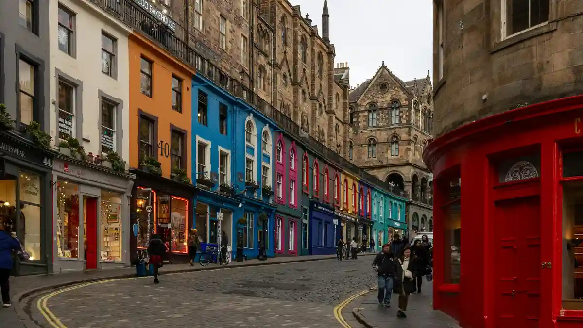 Vibrant street with colorful shopfronts in shades of orange, blue, pink, and red. Stone buildings and cobblestone road enhance the historic feel.