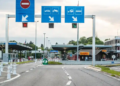 Border checkpoint with lanes for different vehicle types, marked by signs above; a closed lane and open lane indicated. Booths and greenery in the background.